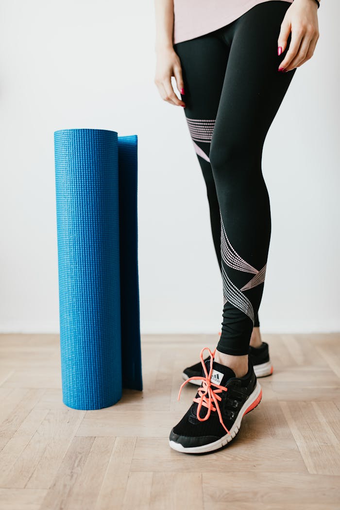 A woman in leggings and sneakers standing with a yoga mat indoors.